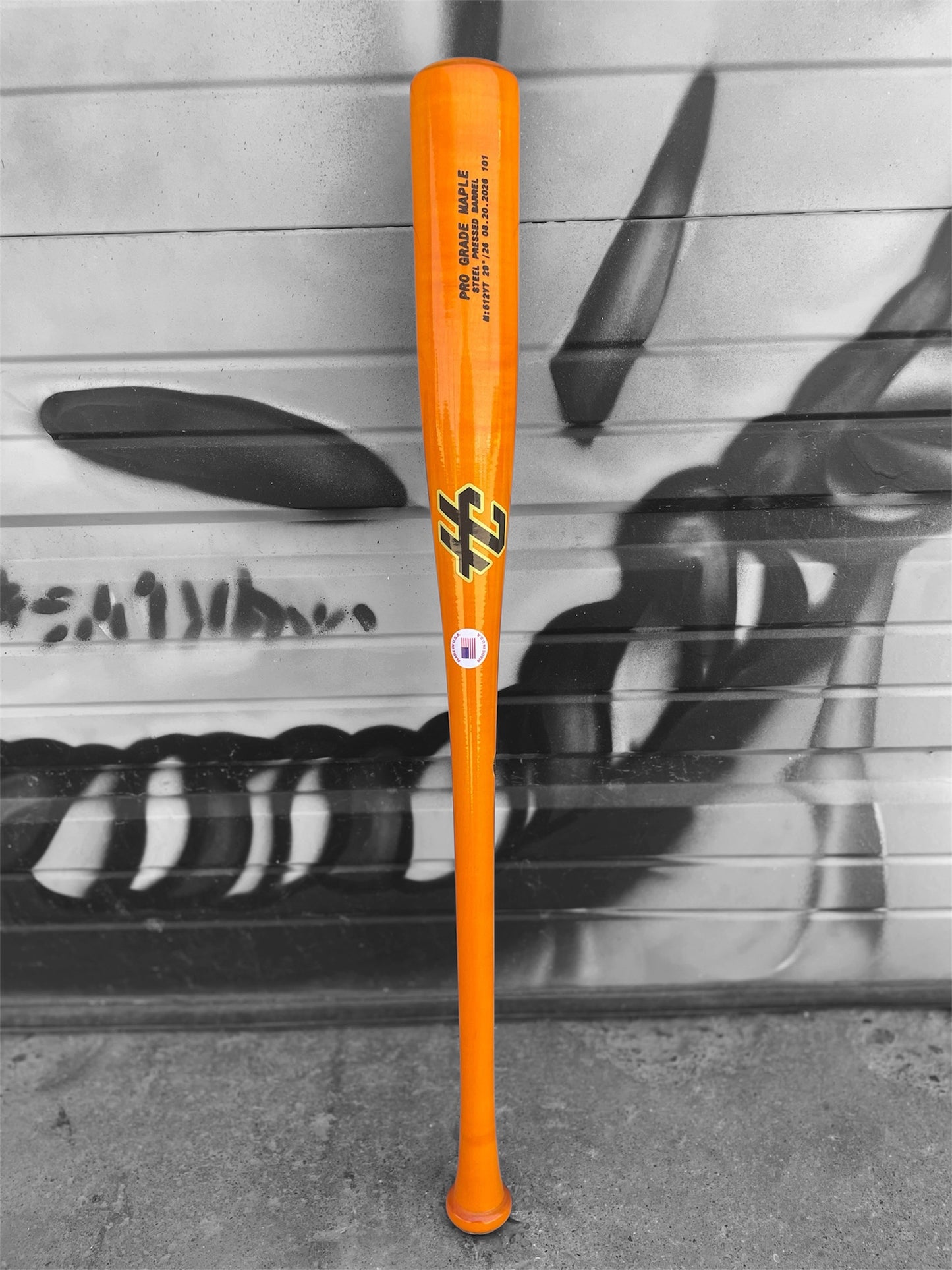 Full-length orange HC Bats 512YT maple baseball bat standing vertically in front of a black and white graffiti wall.
