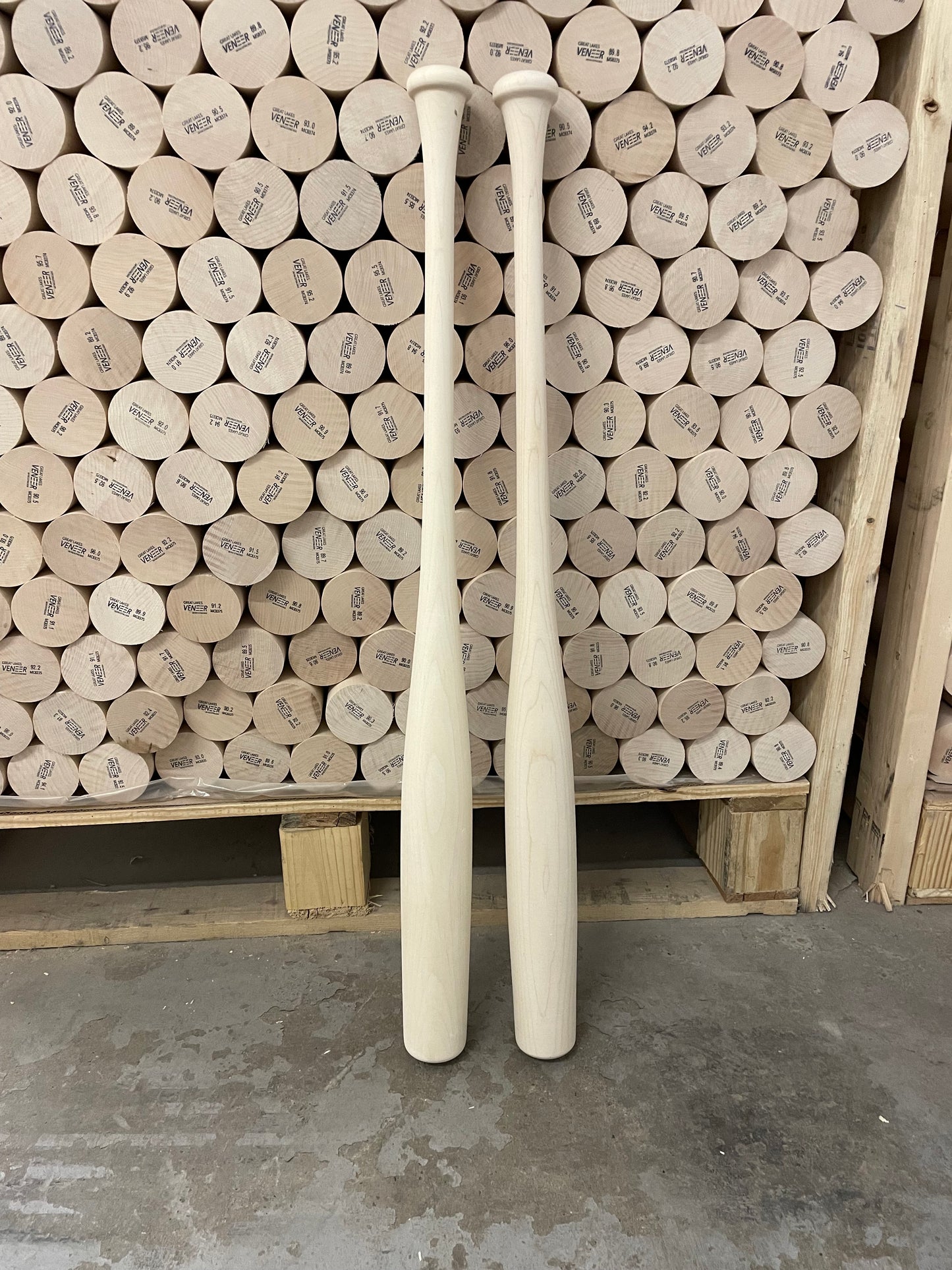 Two wooden baseball bats standing upright against a wall of baseballs, with the bats displaying a natural wood color.
