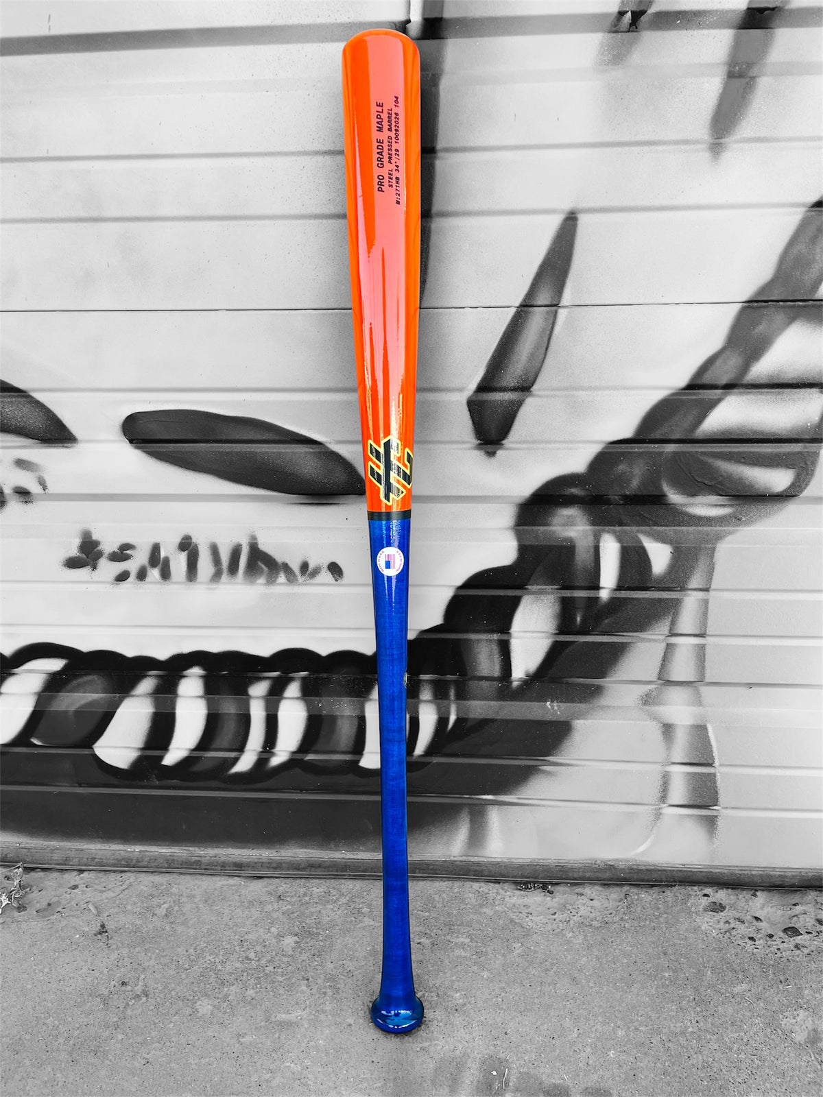 HC Bats 271HB 34-inch Pro Grade Maple baseball bat with royal blue handle and neon orange barrel standing upright against a black and white graffiti-covered garage door.