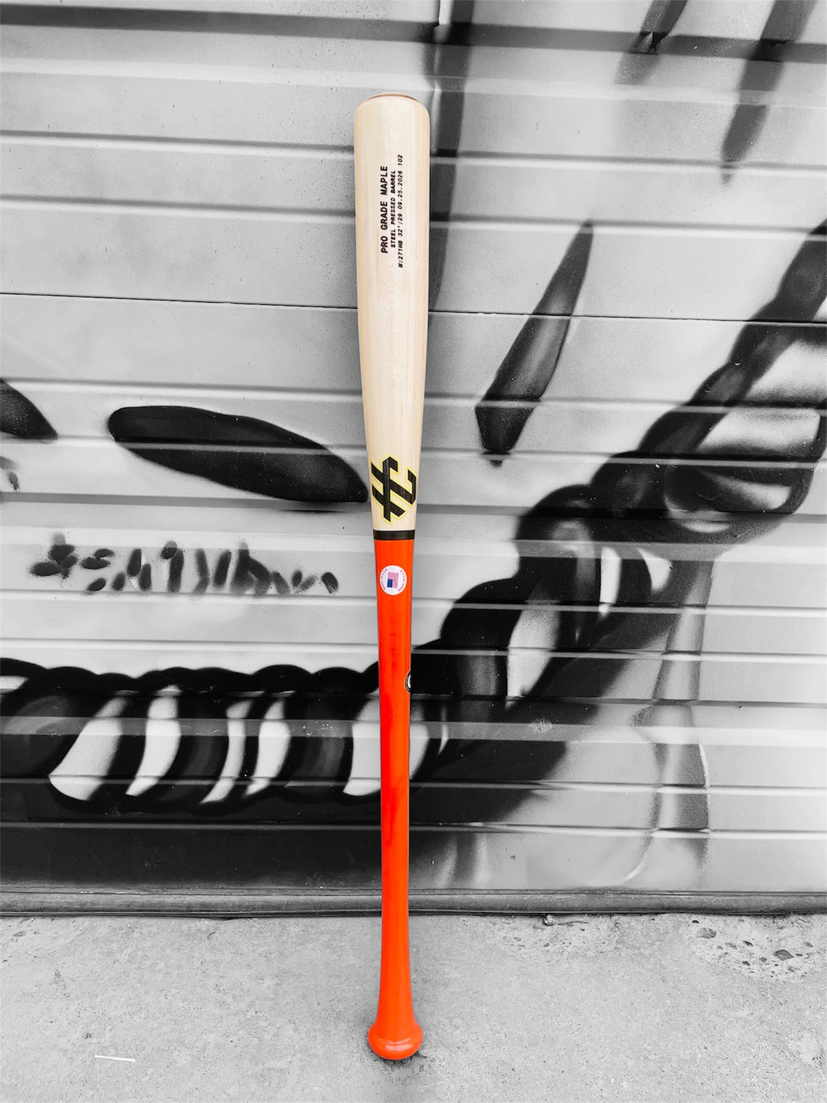 HC Bats Pro Grade Maple 271HB 32-inch 29 oz baseball bat with bright orange handle and natural maple barrel standing upright in front of a black and white graffiti wall.