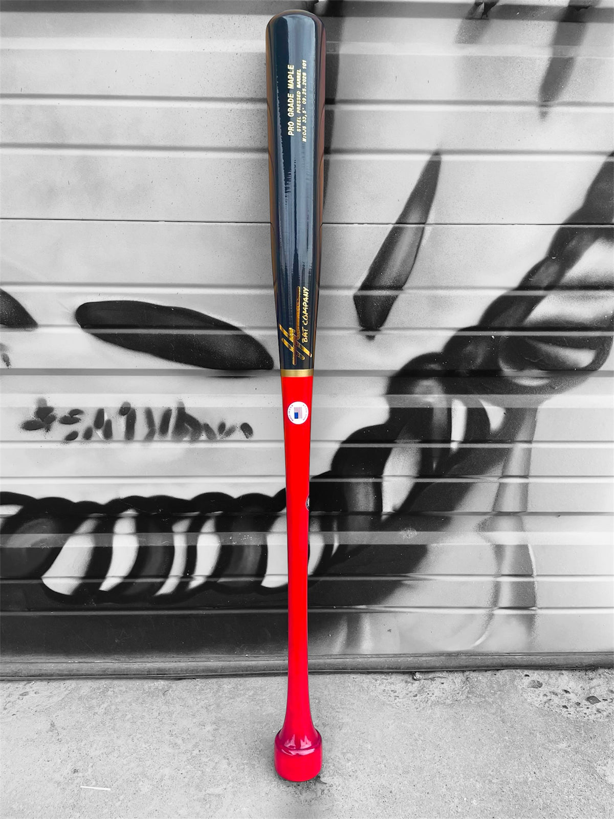 HC Bats CJ5 33.5-inch Pro Grade Maple baseball bat with red handle, hockey puck knob and dark navy-black barrel standing upright in front of a black and white graffiti wall.